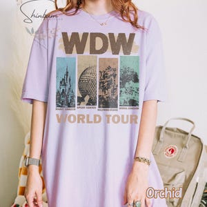 May include: A lavender t-shirt with a vintage-style graphic of the Walt Disney World logo and the text "World Tour". The graphic features the four theme parks: Magic Kingdom, Epcot Center, Hollywood Studios, and Animal Kingdom.
