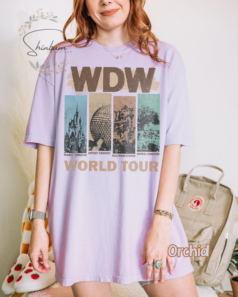 May include: A lavender t-shirt with a vintage-style graphic of the Walt Disney World logo and the text "World Tour". The graphic features the four theme parks: Magic Kingdom, Epcot Center, Hollywood Studios, and Animal Kingdom.