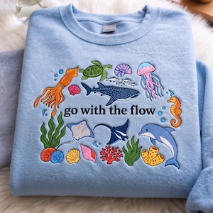 May include: A light blue crewneck sweatshirt featuring an embroidered design of various sea creatures including a whale shark, sea turtle, jellyfish, seahorse, dolphin, squid, and rays, with the text "go with the flow" centred in black lettering.
