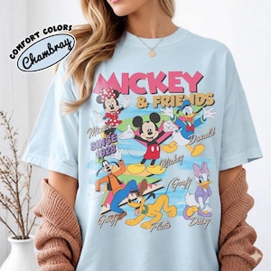 Peut inclure: T-shirt bleu clair avec un motif de Mickey Mouse et ses amis. Le graphisme comprend Mickey, Minnie, Donald, Dingo, Pluto et Daisy. Le texte sur le t-shirt indique "Mickey & Friends" et "Since 1928". Le t-shirt porte le texte "Comfort Colors Chambray" dans un cercle.