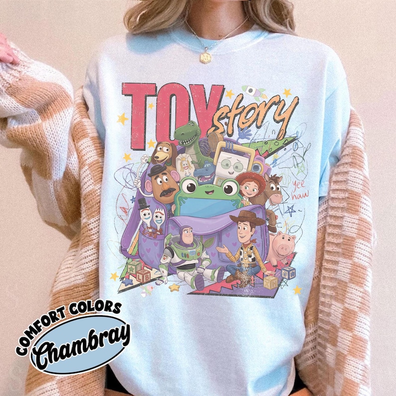 Op de afbeelding: Een lichtblauw sweatshirt met ronde hals en een grafische print van Toy Story-personages, waaronder Woody, Buzz Lightyear en Mr. Potato Head. Het ontwerp bevat de tekst TOY story en yee haw, omringd door kleurrijke sterren en krabbels.