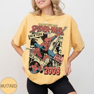 May include: Mustard-coloured t-shirt with a vintage Spider-Man graphic. The design includes the words "Brand New Day," "New York," and "Swing Into Action," with the year "2008." The graphic depicts Spider-Man in action.