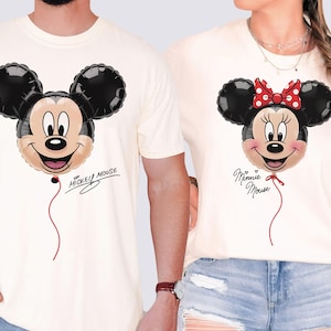 K&ouml;nnte beinhalten: Cremefarbene T-Shirts mit Mickey- und Minnie-Mouse-Ballon-Designs. Mickeys Ballon hat einen schwarzen Kopf und Ohren, ein beiges Gesicht und den Text "Mickey Mouse". Minnies Ballon hat eine rote Schleife und den Text "Minnie Mouse".