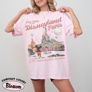 May include: A light pink t-shirt with a graphic of Disneyland Paris. The graphic includes the Disneyland Paris castle, Mickey Mouse, and the text "Paris, France Est. 1992" and "The happiest place on earth."