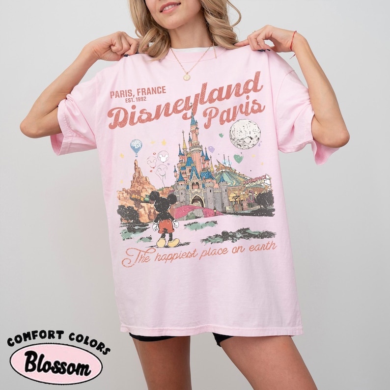 May include: A light pink t-shirt with a graphic of Disneyland Paris. The graphic includes the Disneyland Paris castle, Mickey Mouse, and the text "Paris, France Est. 1992" and "The happiest place on earth."