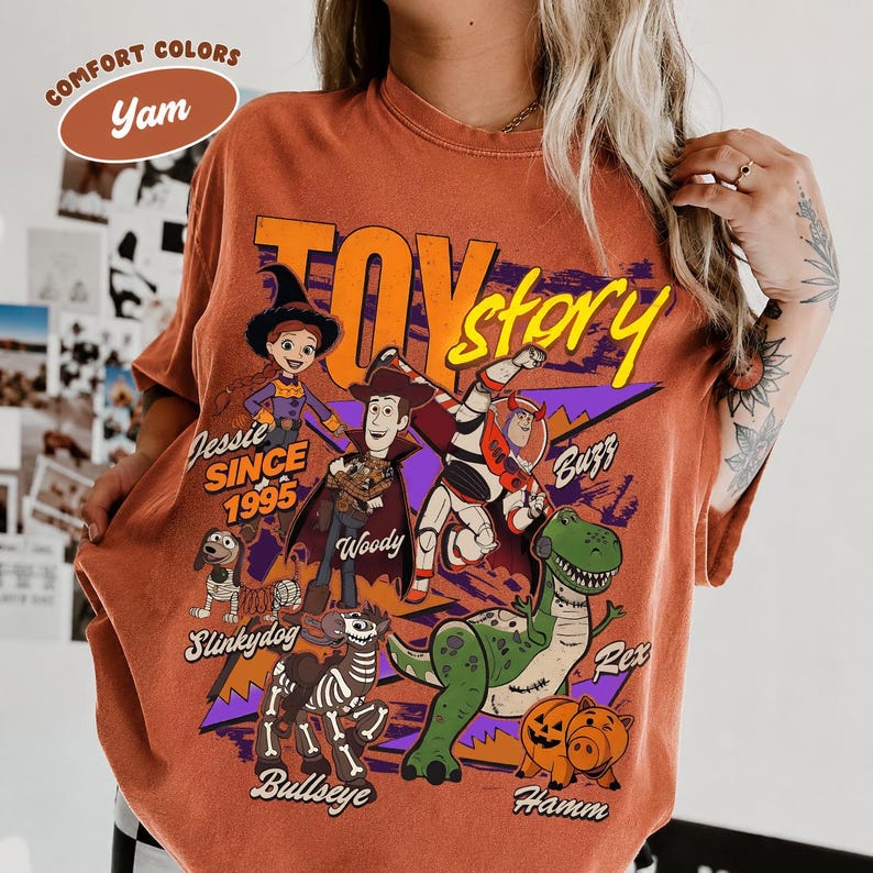 May include: Orange-colored t-shirt featuring characters from the animated film "Toy Story." The design includes Jessie, Woody, Buzz Lightyear, Rex, Slinkydog, Bullseye, and Hamm. The text "Toy Story" and "Since 1995" are also visible.