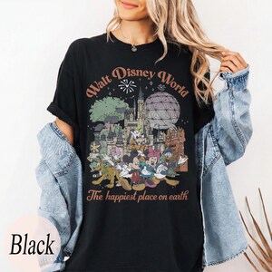 May include: Black t-shirt featuring a Walt Disney World graphic with various cartoon characters and park landmarks. The text "The happiest place on earth" is below the graphic. The shirt is black with the word "Black" at the bottom.