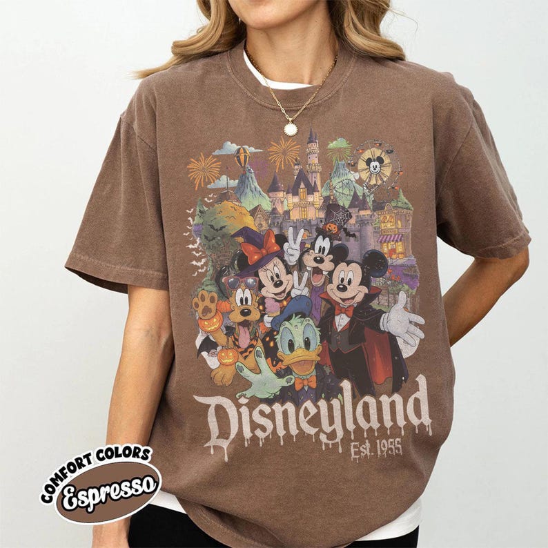 May include: A brown short-sleeve t-shirt featuring a graphic of Disney characters in Halloween costumes in front of a castle. The text reads Disneyland Est. 1955. A small circular label in the corner displays the text COMFORT COLORS Espresso.