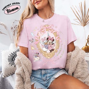 May include: A light pink t-shirt featuring a framed illustration of Minnie Mouse and Daisy Duck. The design includes bows, stars, and the text "Comfort Colors Blossom". The shirt is paired with denim shorts.