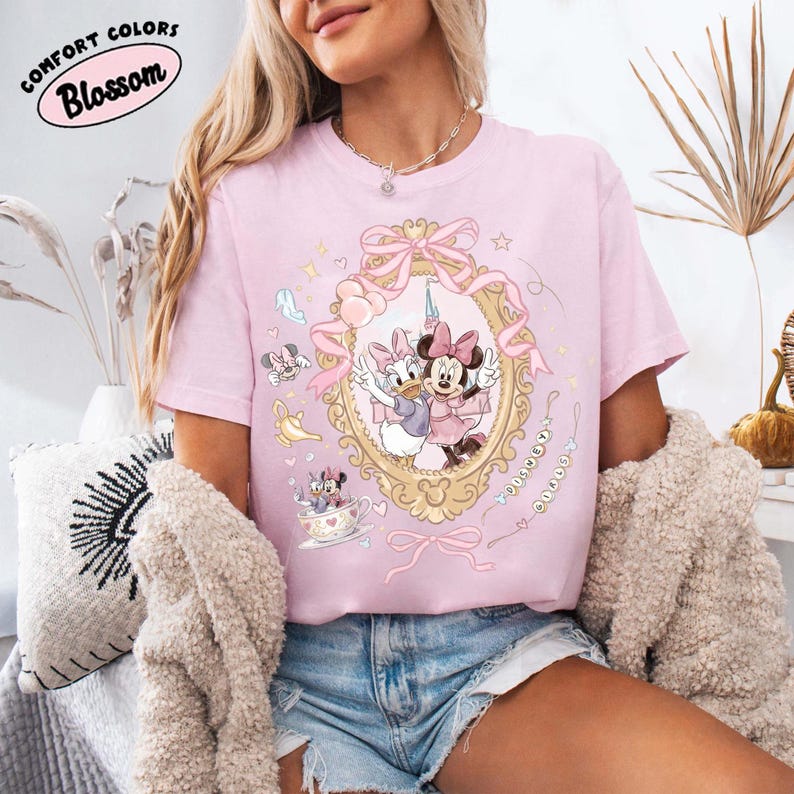 May include: A light pink t-shirt featuring a framed illustration of Minnie Mouse and Daisy Duck. The design includes bows, stars, and the text "Comfort Colors Blossom". The shirt is paired with denim shorts.