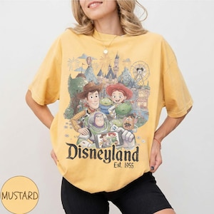May include: A mustard yellow t-shirt featuring a graphic print of Toy Story characters including Woody, Buzz Lightyear, Jessie, Rex, and Mr. Potato Head taking a selfie in front of Disneyland. The text Disneyland Est. 1955 is printed below.