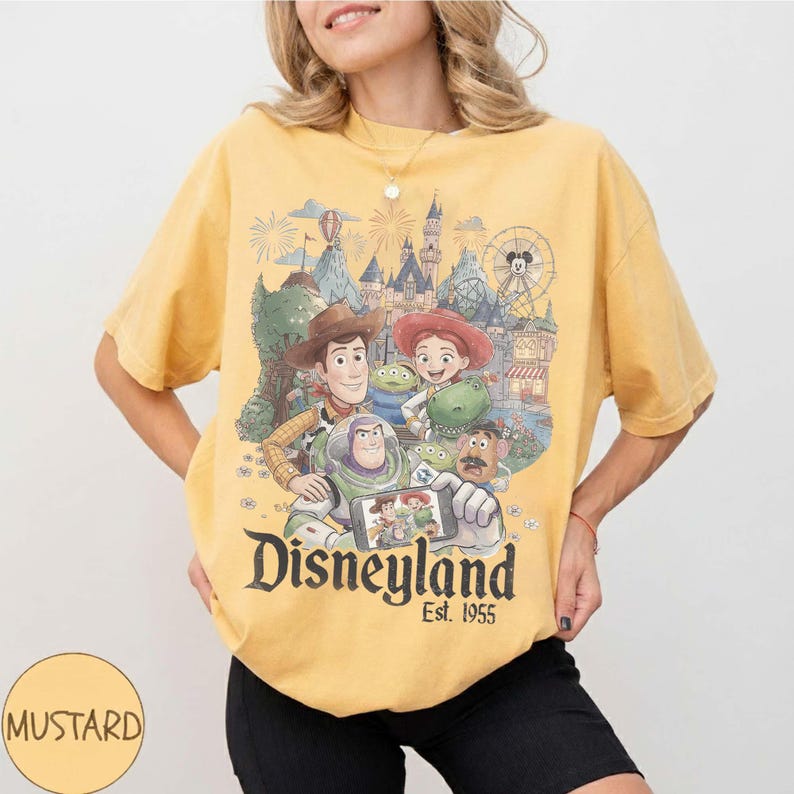 May include: A mustard yellow t-shirt featuring a graphic print of Toy Story characters including Woody, Buzz Lightyear, Jessie, Rex, and Mr. Potato Head taking a selfie in front of Disneyland. The text Disneyland Est. 1955 is printed below.