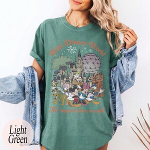 May include: Light green t-shirt featuring a graphic of Disney World characters and landmarks. The design includes Mickey Mouse, Minnie Mouse, Donald Duck, and Goofy, with the text "Walt Disney World" and "The happiest place on earth."
