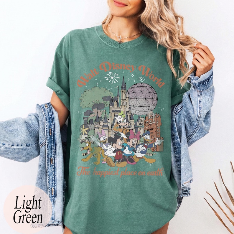 May include: Light green t-shirt featuring a graphic of Disney World characters and landmarks. The design includes Mickey Mouse, Minnie Mouse, Donald Duck, and Goofy, with the text "Walt Disney World" and "The happiest place on earth."