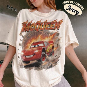 Może przedstawiać: Kremowa koszulka z grafiką Zygzaka McQueena z filmu Auta. Projekt zawiera napis "McQueen" w płomiennych pomarańczowych literach nad czerwonym samochodem wyścigowym. Koszulka ma kr&oacute;tkie rękawy i okrągły dekolt.