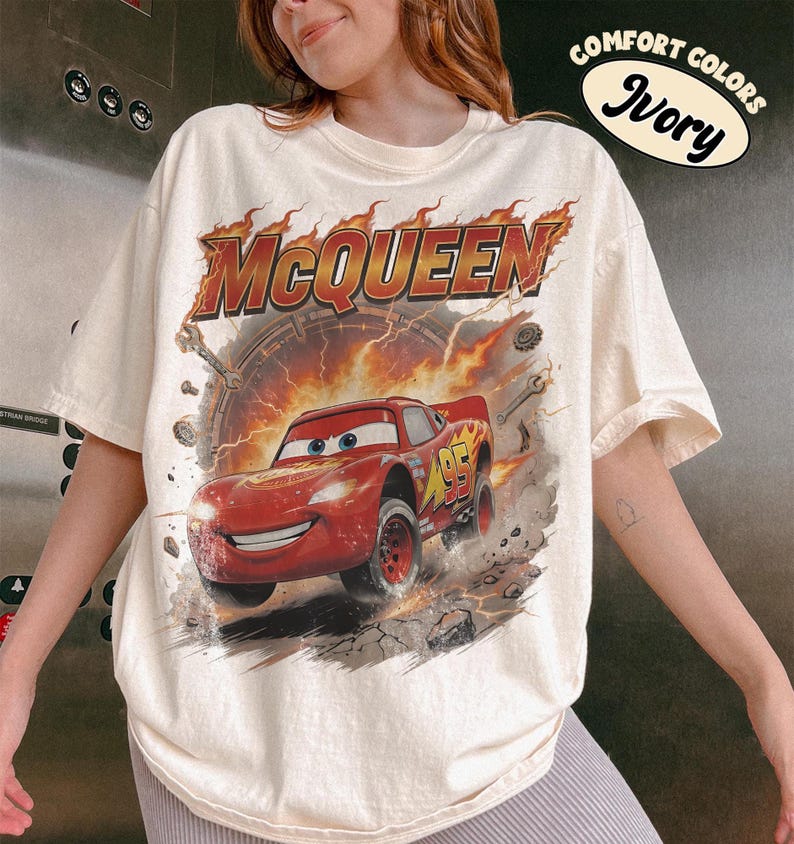 Może przedstawiać: Kremowa koszulka z grafiką Zygzaka McQueena z filmu Auta. Projekt zawiera napis "McQueen" w płomiennych pomarańczowych literach nad czerwonym samochodem wyścigowym. Koszulka ma kr&oacute;tkie rękawy i okrągły dekolt.