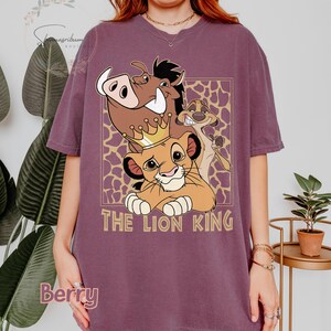May include: Berry-colored t-shirt featuring a graphic of Simba, Timon, and Pumbaa from The Lion King. The design includes a gold crown and the words "THE LION KING" in a gold font. The background has a giraffe print.