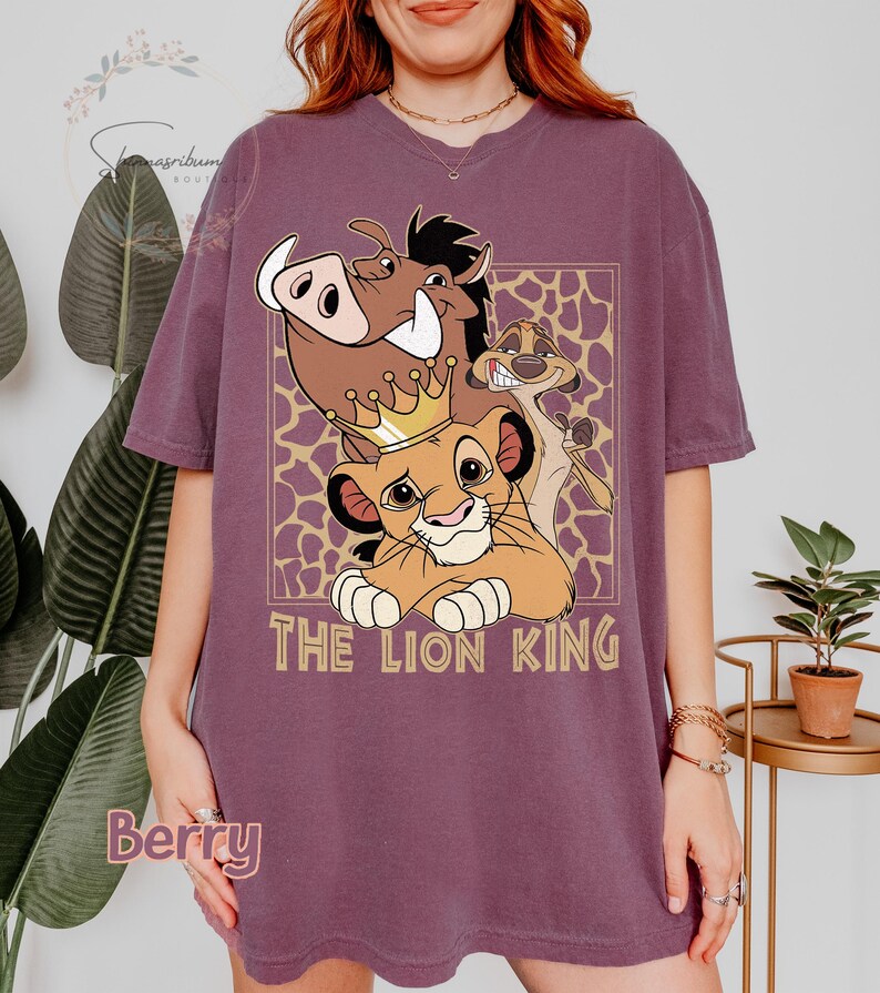 May include: Berry-colored t-shirt featuring a graphic of Simba, Timon, and Pumbaa from The Lion King. The design includes a gold crown and the words "THE LION KING" in a gold font. The background has a giraffe print.