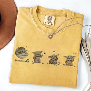 Puede incluir: Camiseta de color amarillo mostaza con un gr&aacute;fico de un personaje de una popular serie de ciencia ficci&oacute;n. El gr&aacute;fico muestra al personaje en diferentes poses con una rana y una taza. La camiseta tiene el texto "Comfort Colors" e informaci&oacute;n de tallas.