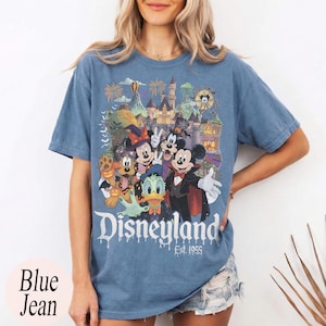 May include: A blue jean colored t-shirt featuring a graphic print of Disney characters including Mickey Mouse, Minnie Mouse, Donald Duck, and Pluto in Halloween costumes in front of a castle with the text Disneyland Est. 1955 printed below.