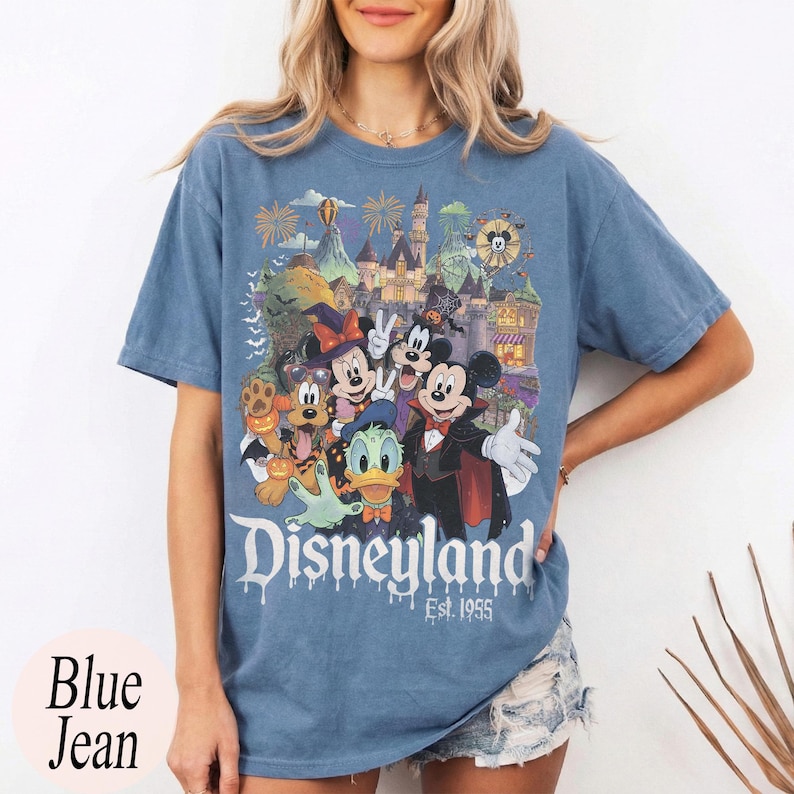 May include: A blue jean colored t-shirt featuring a graphic print of Disney characters including Mickey Mouse, Minnie Mouse, Donald Duck, and Pluto in Halloween costumes in front of a castle with the text Disneyland Est. 1955 printed below.