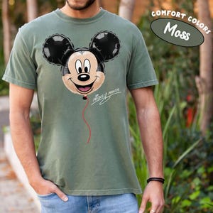 K&ouml;nnte beinhalten: Moosgr&uuml;nes T-Shirt mit einem Mickey Mouse Ballon-Design. Der Ballon ist schwarz und beige mit einem roten Faden, und die Worte "Mickey Mouse" sind in Schrift geschrieben. Das T-Shirt ist mit "Comfort Colors" gekennzeichnet.