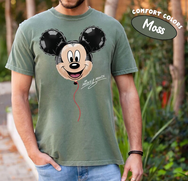 K&ouml;nnte beinhalten: Moosgr&uuml;nes T-Shirt mit einem Mickey Mouse Ballon-Design. Der Ballon ist schwarz und beige mit einem roten Faden, und die Worte "Mickey Mouse" sind in Schrift geschrieben. Das T-Shirt ist mit "Comfort Colors" gekennzeichnet.