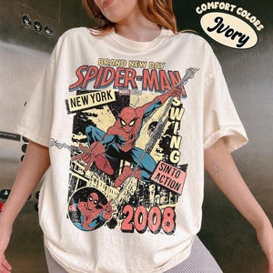 May include: Ivory t-shirt with a Spider-Man graphic. The design includes the words "BRAND NEW DAY," "SPIDER-MAN," "NEW YORK," and "2008." The graphic depicts Spider-Man in action, with the words "SWING" and "SIN TO ACTION."
