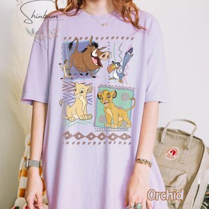 May include: Orchid-colored t-shirt featuring a graphic of characters from The Lion King. The design includes Simba, Timon, Pumbaa, and Zazu in colorful squares. The shirt has short sleeves and a relaxed fit.