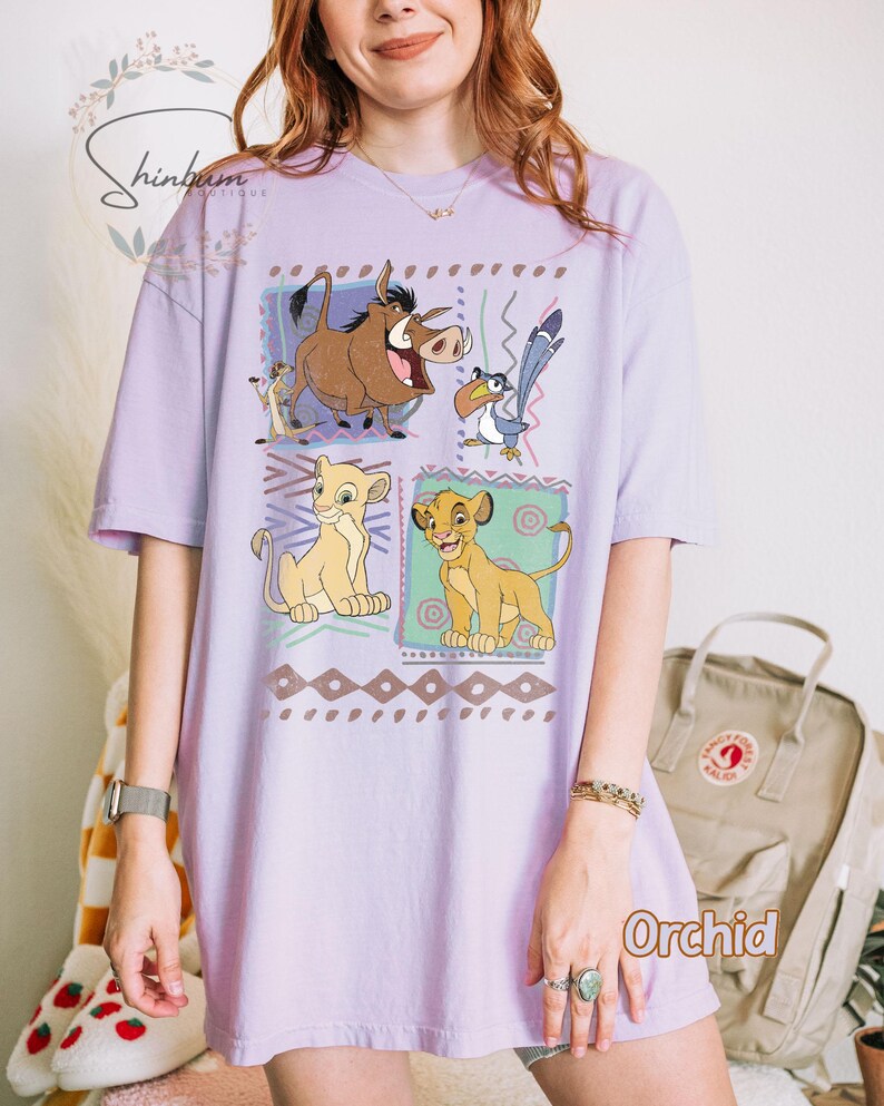 May include: Orchid-colored t-shirt featuring a graphic of characters from The Lion King. The design includes Simba, Timon, Pumbaa, and Zazu in colorful squares. The shirt has short sleeves and a relaxed fit.