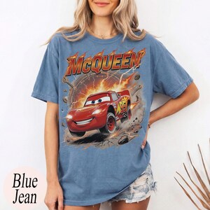 Może przedstawiać: Niebieska koszulka dżinsowa z grafiką Zygzaka McQueena z filmu Auta. Grafika zawiera słowo "McQueen" w płomiennej czcionce i numer 95 na samochodzie. Słowa "Blue Jean" są wydrukowane na dole.