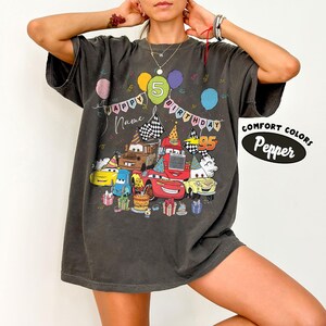 K&ouml;nnte beinhalten: Dunkelgraues T-Shirt mit einer bunten Grafik von Zeichentrickautos, Luftballons und Geburtstagskuchen. Der Text lautet "Happy Birthday" und "Name" &uuml;ber den Autos, mit der Zahl "5" auf einem Ballon. Das Shirt hat auch die Worte "Comfort Colors" und "Pepper".