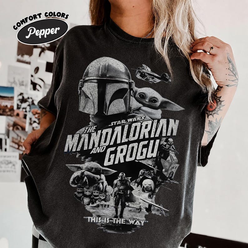 Op de afbeelding: Donkergrijs t-shirt met een afbeelding van The Mandalorian en Grogu uit Star Wars. Het ontwerp bevat de helm van de Mandalorian, Grogu, en de tekst "THE MANDALORIAN AND GROGU" en "THIS IS THE WAY". Het shirt heeft een vintage, distressed look.