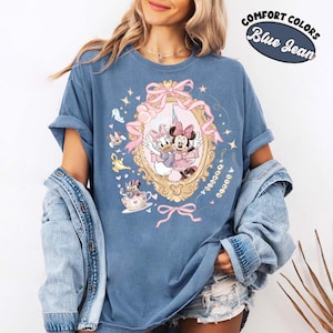 May include: Blue jean-colored t-shirt featuring a framed illustration of Minnie and Daisy Duck. The illustration is surrounded by pink bows and stars. The shirt has the words "Comfort Colors" and "Blue Jean" in a circular design.