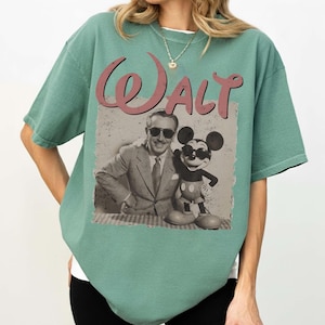 May include: Sage green t-shirt featuring a vintage-style graphic of Walt Disney with Mickey Mouse. The graphic includes the word "Walt" in a pink, cursive font. The shirt is a relaxed fit.