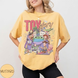Op de afbeelding: Een mosterdgeel T-shirt met korte mouwen en een grote grafische print van Toy Story-personages, waaronder Woody, Buzz Lightyear en Rex. Het ontwerp bevat de tekst "TOY story" in dikke rode en zwarte letters met kleurrijke krabbels.