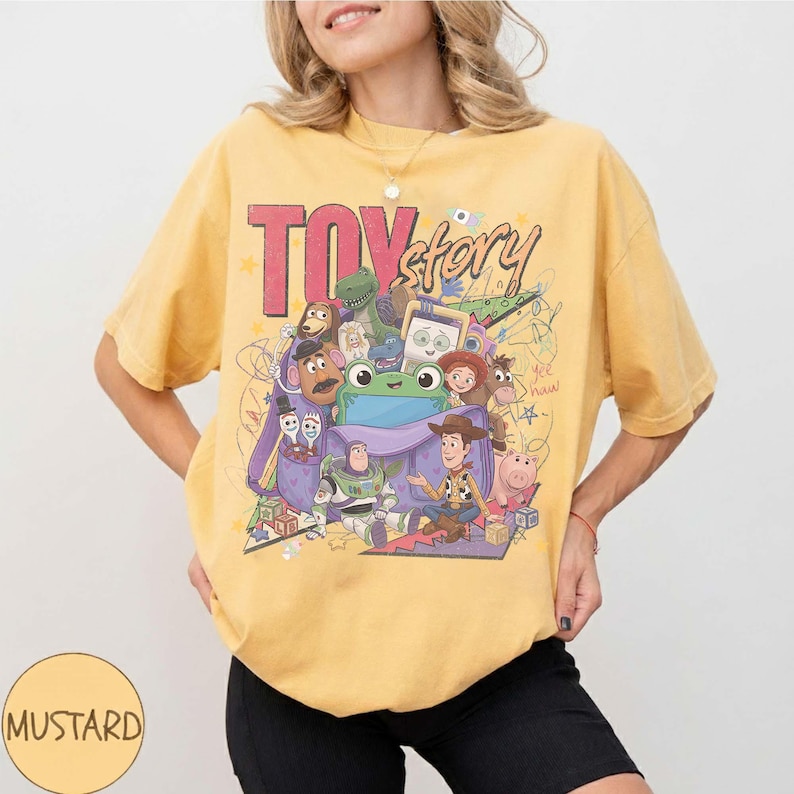 Op de afbeelding: Een mosterdgeel T-shirt met korte mouwen en een grote grafische print van Toy Story-personages, waaronder Woody, Buzz Lightyear en Rex. Het ontwerp bevat de tekst "TOY story" in dikke rode en zwarte letters met kleurrijke krabbels.