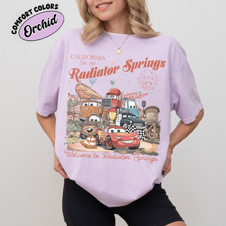 以下が含まれることがあります： 映画「カーズ」のキャラクターのグラフィックが特徴の薄紫色のTシャツ。「California Est. 2012 Radiator Springs」と「Welcome to Radiator Springs」の文字入り。シャツには「Comfort Colors」と「Orchid」の文字も円で囲まれています。