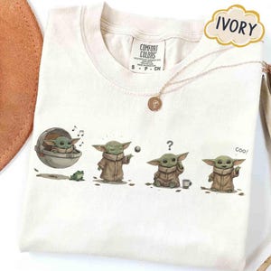 Puede incluir: Camiseta de color marfil con un gr&aacute;fico de un personaje verde con una capa marr&oacute;n, en varias poses. La palabra "IVORY" est&aacute; en una burbuja con forma de nube. Tambi&eacute;n se ve un collar dorado.
