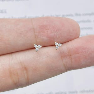 May include: A pair of gold stud earrings, each featuring three small, clear, round-cut stones arranged in a triangular cluster. The earrings are delicate and minimalist in design, showcasing a bright metallic finish and sparkling stones.