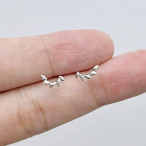 Puede incluir: Un par de pequeños pendientes de botón de plata, cada uno con forma de rama curva con tres segmentos distintos y pulidos en forma de hoja. Los pendientes presentan un acabado metálico reflectante y un diseño de silueta arqueada.
