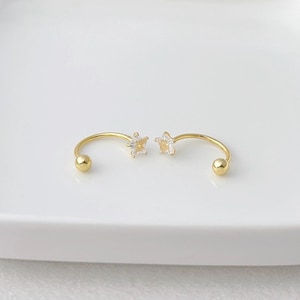 May include: A pair of gold-colored, open-hoop earrings. Each earring features a small, clear, star-shaped gem at the center of the hoop. The ends of the hoops have a small, gold-colored ball. The earrings are displayed on a white surface.