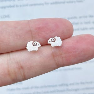 May include: A pair of small, silver sheep-shaped stud earrings. Each earring has a spiral design for the wool and a dot for the eye. The earrings are shown against a light background, ideal for everyday wear.