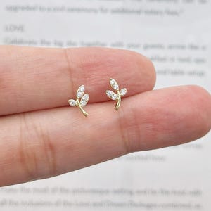 May include: A pair of gold stud earrings shaped like small branches with three clear, sparkling stones set into each leaf. The earrings feature a delicate, curved design that mimics the appearance of a sprig, crafted with a polished gold finish.