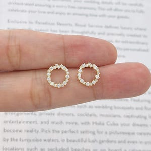 Peut inclure: Une paire de boucles d'oreilles rondes dorées. Chaque boucle d'oreille présente un anneau de petites pierres précieuses transparentes. Les boucles d'oreilles sont tenues entre les doigts sur un fond de texte imprimé.
