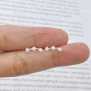 May include: Delicate silver earrings with a floral design. Each earring has a curved bar with small, detailed flower blossoms. The earrings are shown on a person's finger against a blurred background.