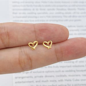 以下が含まれることがあります： 小さくて金色のハート型のスタッドピアス2つ。ピアスはシンプルで繊細なオープンハートのデザインです。明るい色の背景に表示されています。
