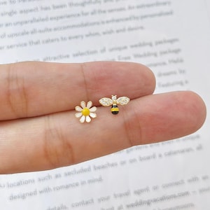 Peut inclure: Une paire de boucles d'oreilles à clous dorées. L'une est une marguerite blanche avec un centre jaune. L'autre est une abeille avec un corps noir et jaune et des ailes en forme de cristal. Les boucles d'oreilles sont petites et délicates.