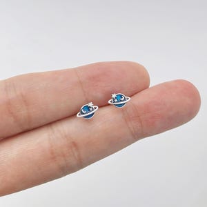 Op de afbeelding: Een paar kleine, zilverkleurige oorstekers in de vorm van een planeet. Elke oorbel heeft een blauw planeetontwerp met een zilveren ring, een maan en een ster. De oorbellen worden op een lichte achtergrond getoond.