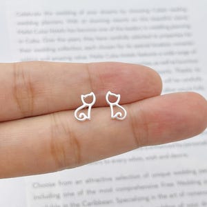 May include: A pair of silver cat-shaped stud earrings. Each earring has a minimalist outline of a sitting cat with a curled tail. The earrings are small and delicate, suitable for everyday wear. The background is a blurred image of text.